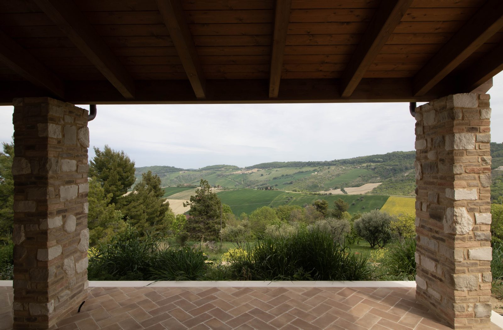 Portico della casa in legno di Monterubbiano con travi di legno a vista e pilastri in pietra che inquadrano le colline